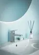 Chromene Armatur von Ideal Standard vor einer blauen Wand, montiert auf einem weißen Waschbecken. Sichtbar sind ein Seifenspender und ein Raumduft.
